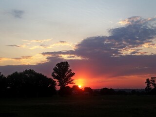 Scenic rural sunset with glowing sun partially hidden by fluffy clouds, bathing fields in warm...