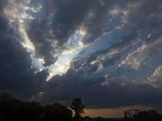 Dramatic sunset over a wide field with dark storm clouds, golden light breaking through the...