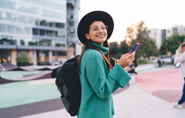 Stylish young female holding smartphone and notebook, wearing backpack and smiling while walking...