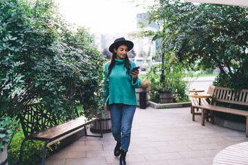 Millennial woman walking down modern sidewalk with smartphone and laptop, smiling while reading...