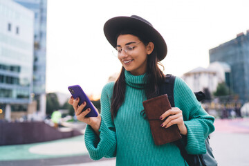 Smiling woman in hat and glasses looking at smartphone while holding planner, showcasing casual...