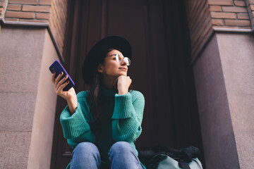 Close-up of woman seated on stairway with phone in hand, chin resting on fist, calm and thoughtful...