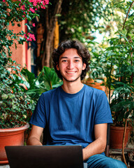 A young man in a blue t-shirt smiles while sitting outdoors with a laptop, surrounded by lush green plants and vibrant flowers.