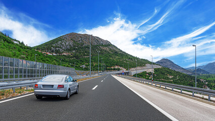 A silver sedan drives along a modern mountain highway under a bright blue sky, showcasing a scenic...