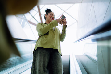 Cheerful woman enjoys scrolling through smartphone while riding an escalator. Lost in social...