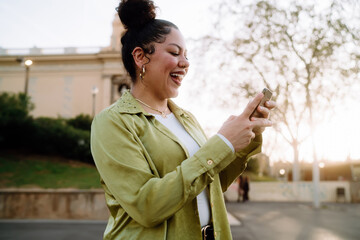 young happy Hispanic woman laughs while using her smartphone outdoors during golden hour. warm...
