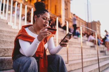 young woman in casual fashion sits on city steps, completing an online transaction. Female checks...