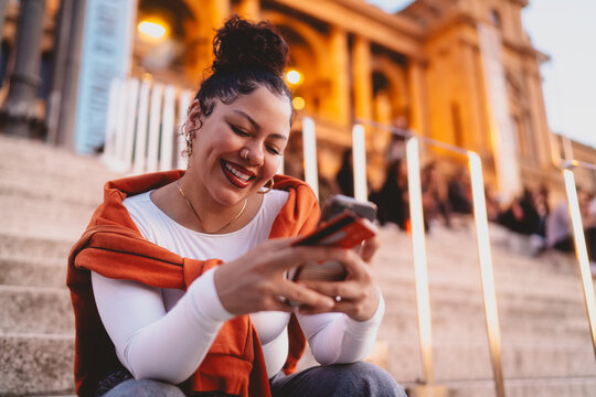 young woman enjoys online shopping while relaxing on historic city steps. Female holds credit card and smartphone, bright smile reflecting satisfaction with digital convenience and financial solutions