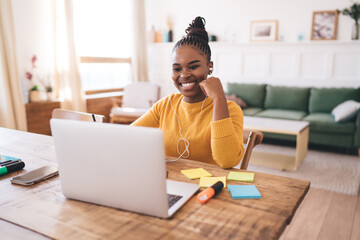 Smiling young Black woman working remotely with laptop and earphones, enjoying positive video call...