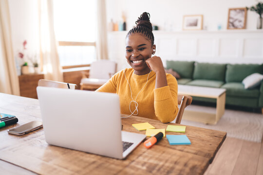 Smiling young Black woman working remotely with laptop and earphones, enjoying positive video call in cozy home interior with sticky notes and stationery on wooden desk