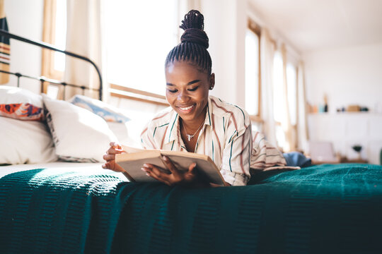 Young woman reading a book on bed, smiling and enjoying relaxing time in bright bedroom, dressed casually in striped shirt and jeans, sunlit room filled with books