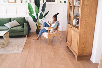Barefoot black woman reading book and drinking coffee in modern armchair, enjoying peaceful moment in cozy bright living room with wooden floors and green sofa