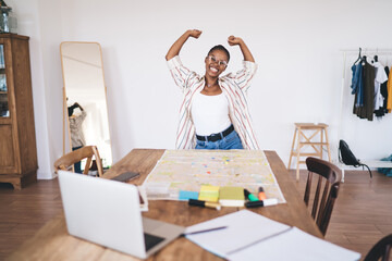 Joyful young Black woman in striped shirt celebrating successful travel planning, raising arms...