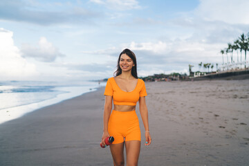 Young brunette walking near sea holding modern speaker, combining fitness, fashion and digital presence in relaxed beach setting.