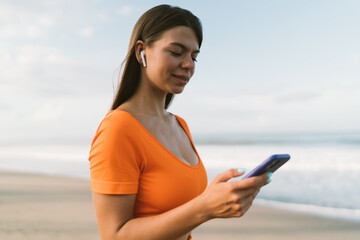 Naklejka premium Young woman in bright outfit focused on smartphone screen while wearing wireless earbuds, symbolizing digital lifestyle, tech immersion and mobile connectivity in outdoor freedom.