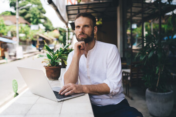 Thoughtful freelancer staring ahead with hand on chin, paused during laptop session. Depicts digital decision-making, remote strategy thinking and modern business reflection outdoors.