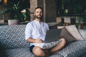 Confident freelancer sitting cross-legged on sofa with laptop, ready to work. Scene of balance between comfort and commitment in self-guided digital career using modern tech tools.