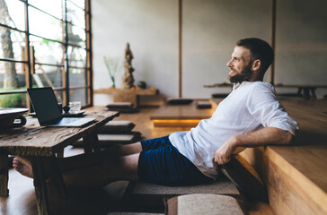 Male freelancer reflecting with calm smile after digital task, symbolizing positive emotions tied to flexible work routines, independence and the modern harmony of life and remote technology.