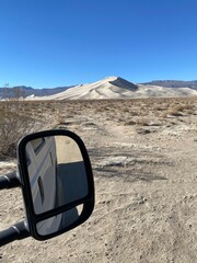 View of a sandy dune landscape with a vehicle mirror reflecting a desert scene under a clear blue...