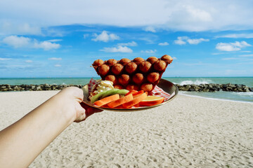 Hand holding freshly made Hong Kong Bubble egg waffles against beach and blue sky background. Hong...