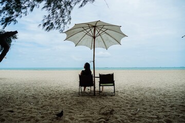 The woman on vacation is sitting in a camping chair under beach umbrella on the beach, enjoying a perfect summer day, sipping coffee, and gazing at the sea in Thailand. Holiday concept.