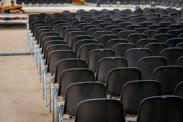 Fototapeta premium Multiple rows of identical black chairs are neatly arranged in an outdoor setting on a dirt surface, creating a uniform and symmetrical pattern.