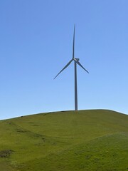 Wind turbine stands tall on green hill under clear blue sky at midday, showcasing renewable energy...