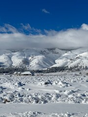 Snow-covered mountains loom over a serene winter landscape with a quaint cabin nestled in the...