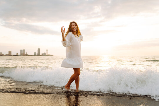 Cheerful woman in white dress enjoys sunset while walking along the seashore. Young woman feels freedom in the open air. Enjoyment, relaxation and weekend concept. - Powered by Adobe