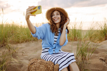 Selfie time! Happy woman on the beach with a phone enjoying the sunset on the beach. Young tourist...