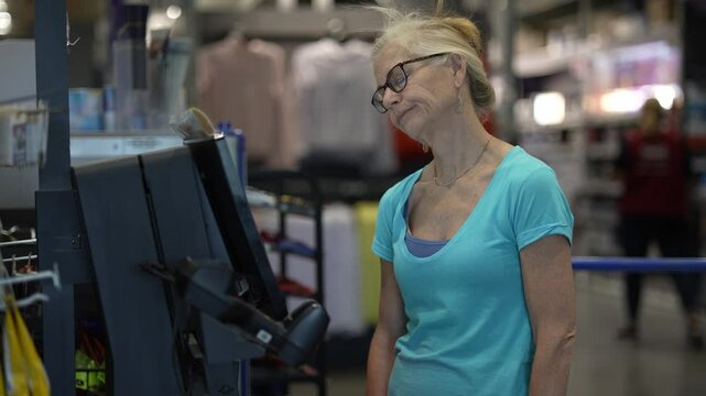 An unhappy, frustrated mature woman stands at the self checkout in a hardware store, looking slightly confused as she processes her purchases with a credit card.
