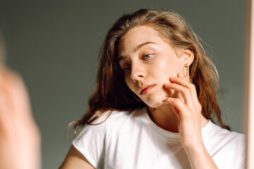 Close-up face of a young woman with rashes looking at the mirror in a cozy room. Beautiful woman...