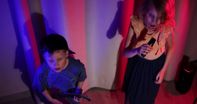 energetic kids stage concert performance at home. boy playing cardboard guitar and girl sing in the microphone. neon lighting