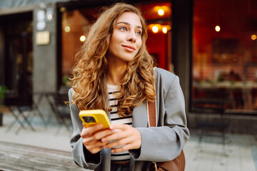 Portrait of a happy woman with a phone in her hands walking along the streets of the city. A stylish woman blogs and texts outdoors. The concept of walking, blogger. Active lifestyle.