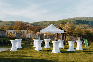 Outdoor event setup with white tables and a tent in a garden setting during daytime	