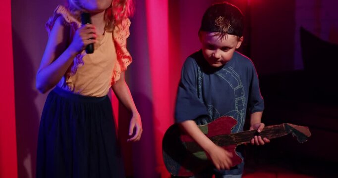 energetic kids stage concert performance at home. boy playing cardboard guitar and girl sing in the microphone. neon lighting