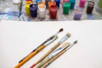 Brushes on a white sheet of paper and jars of multi-colored paint for drawing on the table