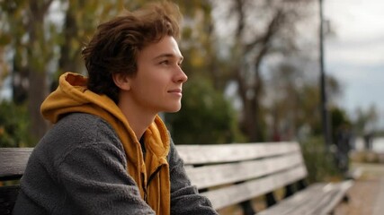 A young man is lost in thought as he sits on a park bench, looking off into the distance