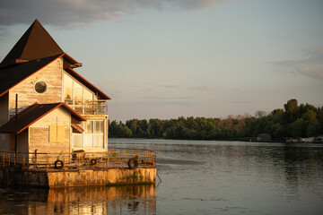 A charming house with distinctive design sits by a peaceful river as sunlight creates a warm glow...