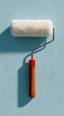 Obraz premium White paint roller with a wooden handle rests against a textured light blue wall, casting a clear shadow.