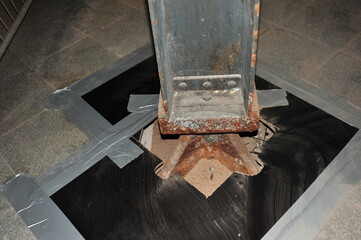 The base of a metal column is bolted to a concrete foundation. The metal surface is rusty, with...