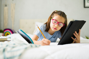 Smart girl doing homework and using tablet on bed