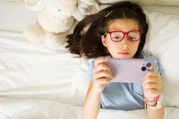 Young girl wearing glasses playing with smartphone in bed