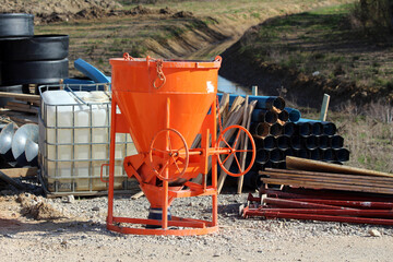 A vivid orange concrete bucket with dual handwheels stands on gravel, surrounded by pipes, wooden...
