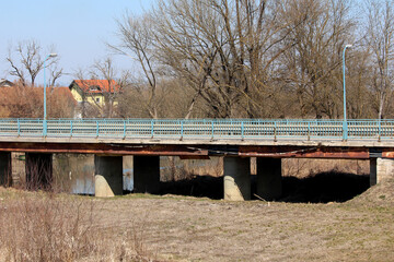 A concrete bridge with blue railings spans a small river, showing visible rust, sagging beams, and...