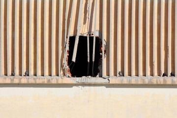 A damaged exterior wall with vertical concrete beams features a large, irregular hole exposing red...