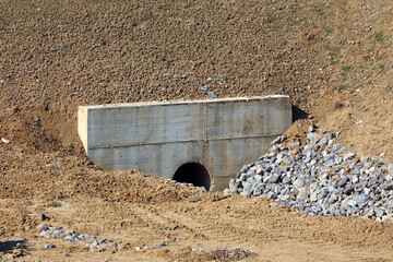 A reinforced concrete culvert pipe emerges from a sloped, eroded earth embankment, surrounded by...