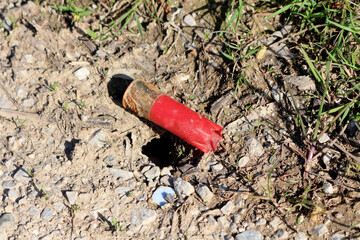 A weathered red shotgun shell casing lies abandoned in the dirt and gravel, partially buried and...