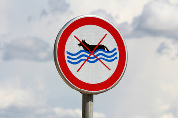A circular prohibition sign features a black dog silhouette swimming in blue waves, crossed out...
