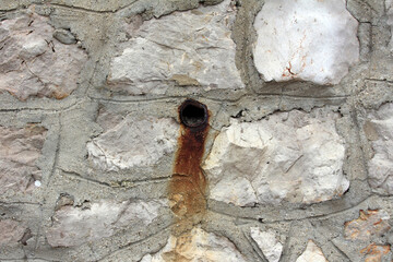 A corroded metal drain pipe juts from a weathered stone wall, leaving a streak of rust down the...